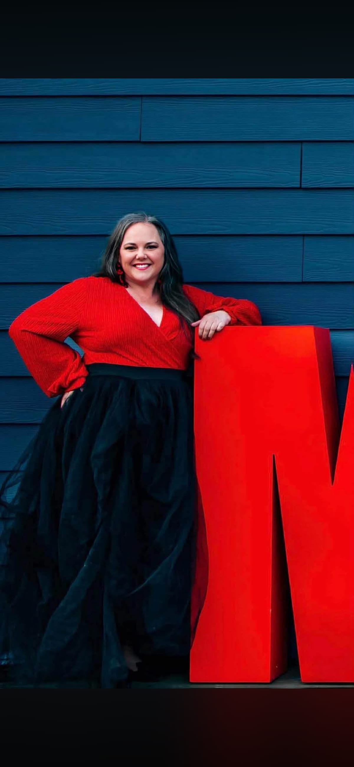 Kim Shelton in a red top and black tulle skirt, leaning on a giant red letter